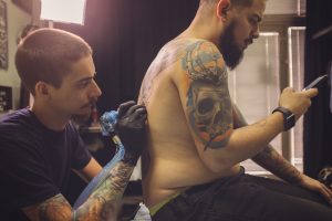 Tattoo artist tattooing his client in his workshop while he using smart phone to help deal with a long tattoo session